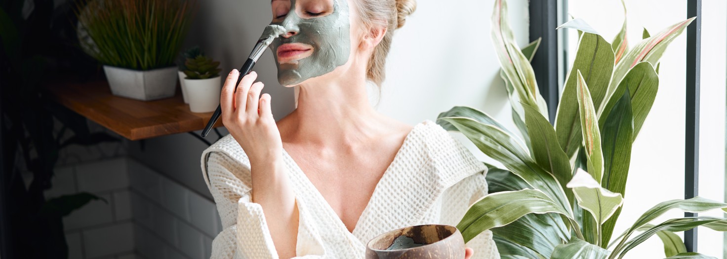 Positive young female in white bathrobe applying purifying clay mask on face with brush from bowl while sitting near window and relaxing during skincare procedure at home
GettyImages-1337589965.jpg